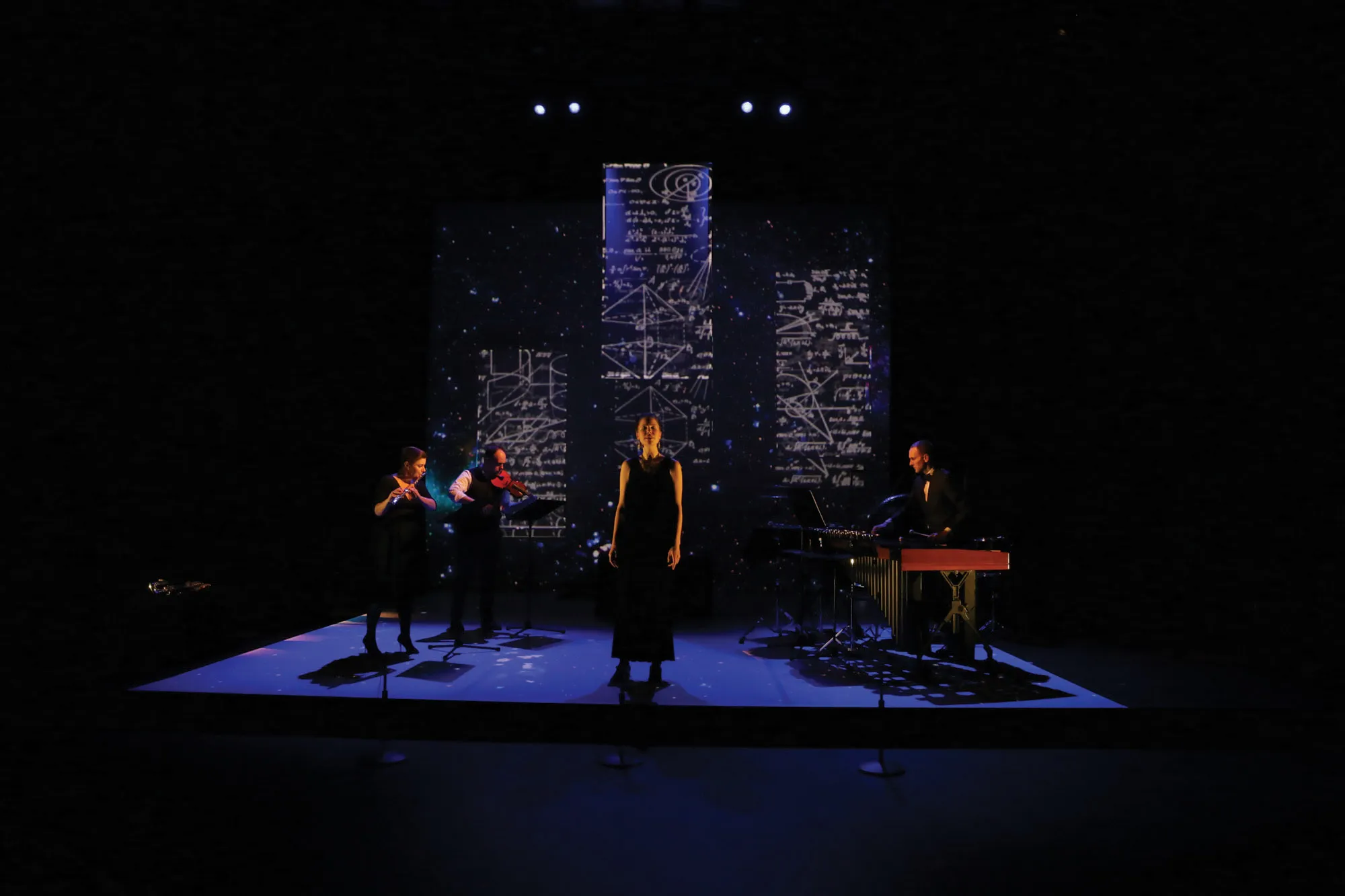A woman standing on a dark stage lit with blue light in front of projections mathematical equations was three musicians play behind her. 
