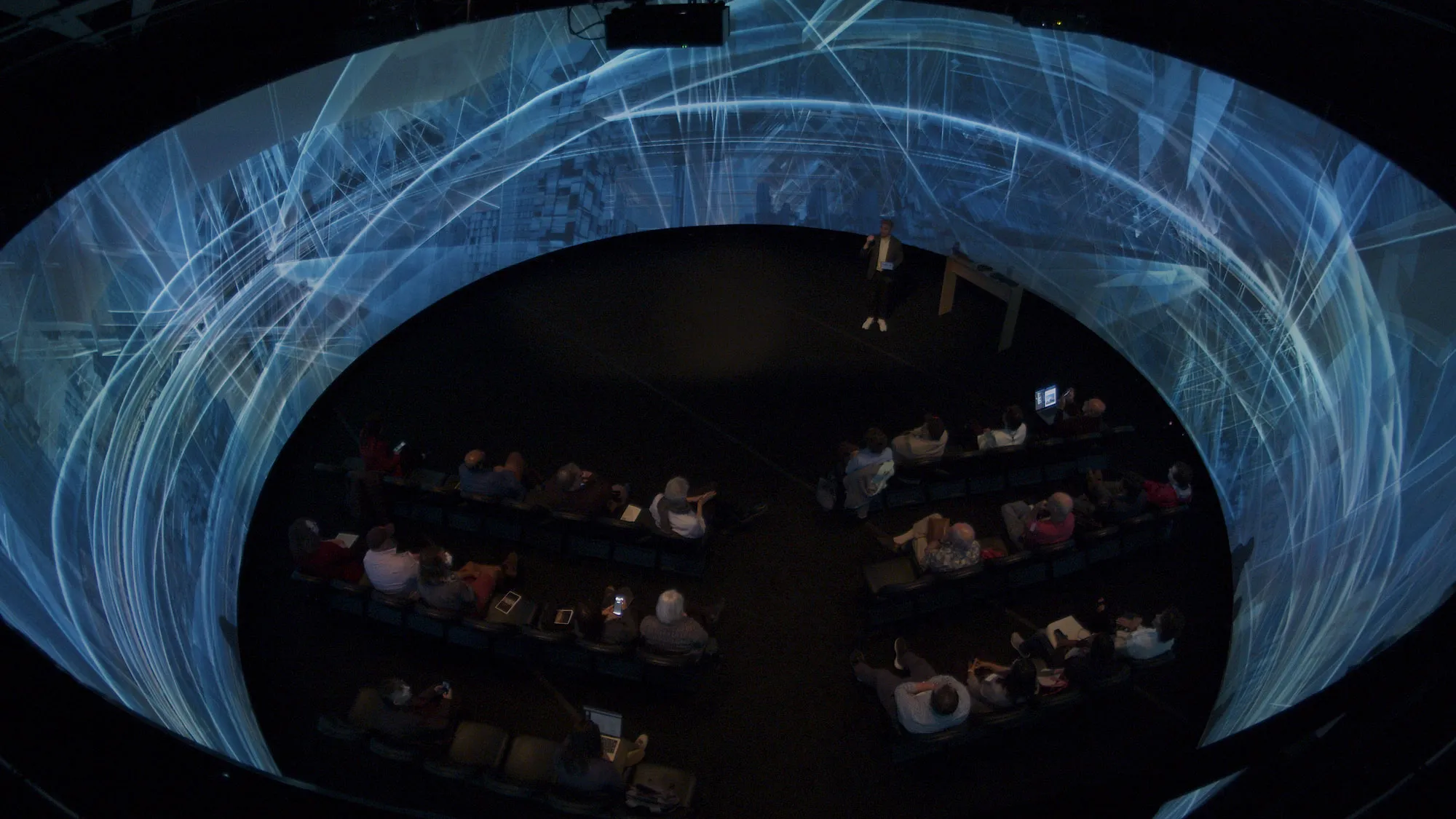 a group of people sit inside a 360 degree panoramic screen listening to a presentation. The screen has complex architectural imagery projected on it.
